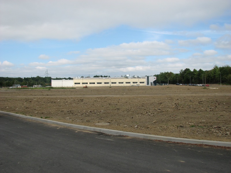 Krajobraz z budynkiem przemysłowym w tle i ugorem na pierwszym planie. Błękitne niebo z chmurami nad polem.